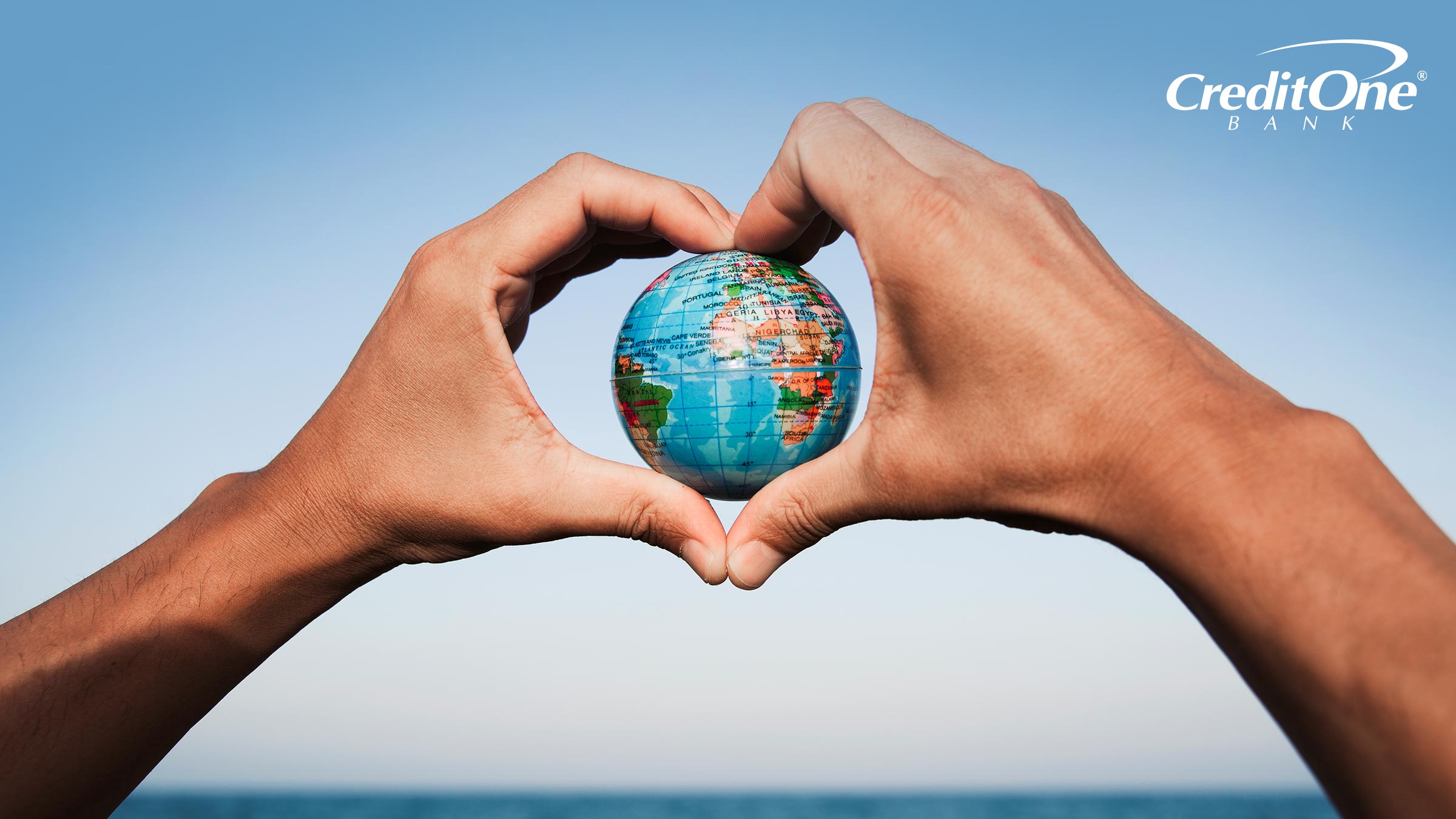 A man’s heart-shaped hands hold a globe in front of the ocean, representing a sustainable vacation