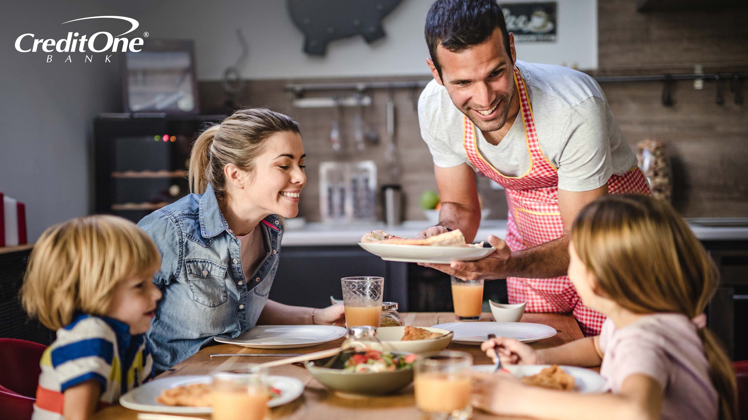 A father brings a plate of food to the dinner table as his wife and two children smile in anticipation. They’ve opted to dine in rather than eat out.