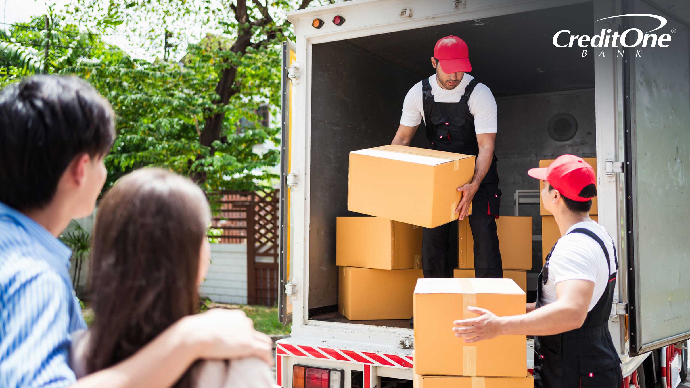A couple watches two professional movers unload boxes from the back of a moving truck. In their situation, professionals may have been the best way to relocate on a budget.