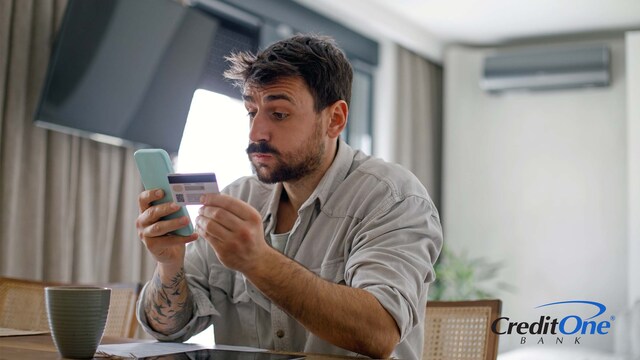 A man holds his phone in one hand and credit card in the other, looking into his phone with a surprised expression. Perhaps this is his first credit card and he narrowly avoided making a mistake.