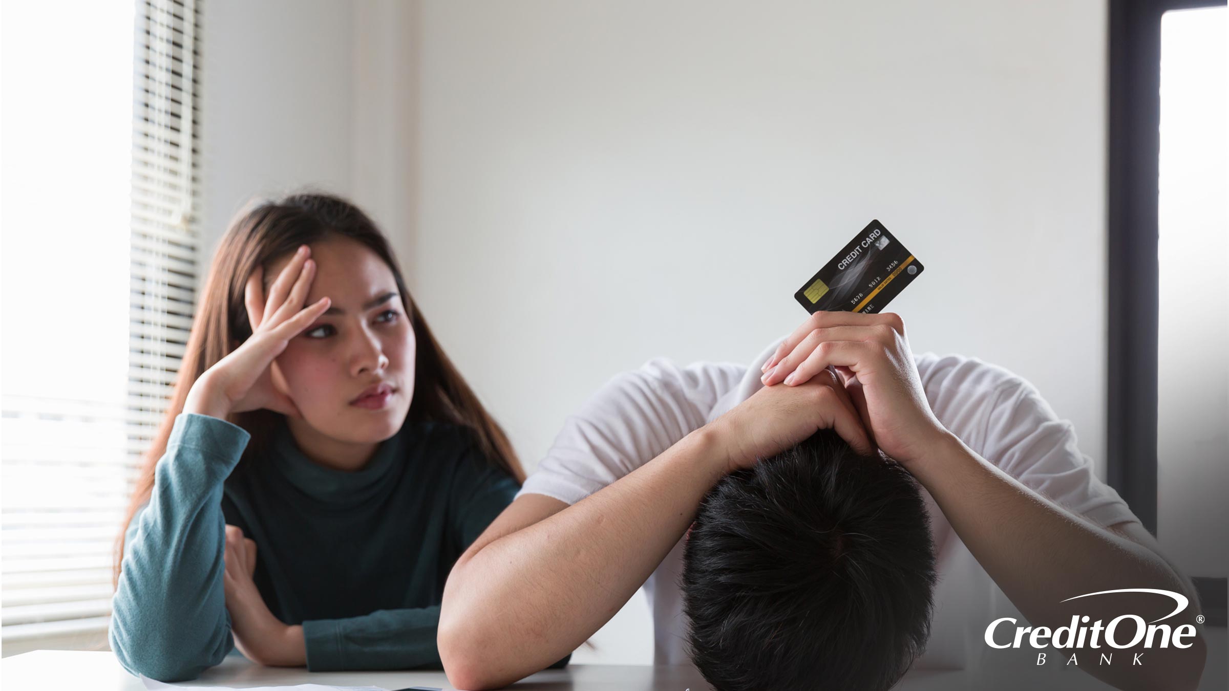 A couple sits at a table, the woman looking at the man with concern and frustration. The man has his face against the tabletop in shame, resting his hands on the back of his head while holding up a credit card. Perhaps he had been hiding his debt from loved ones.