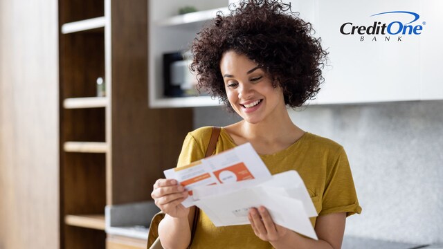 A young woman stands in the kitchen smiling as she reads her newly opened credit card statement, which should include Schumer box information like rates and fees.