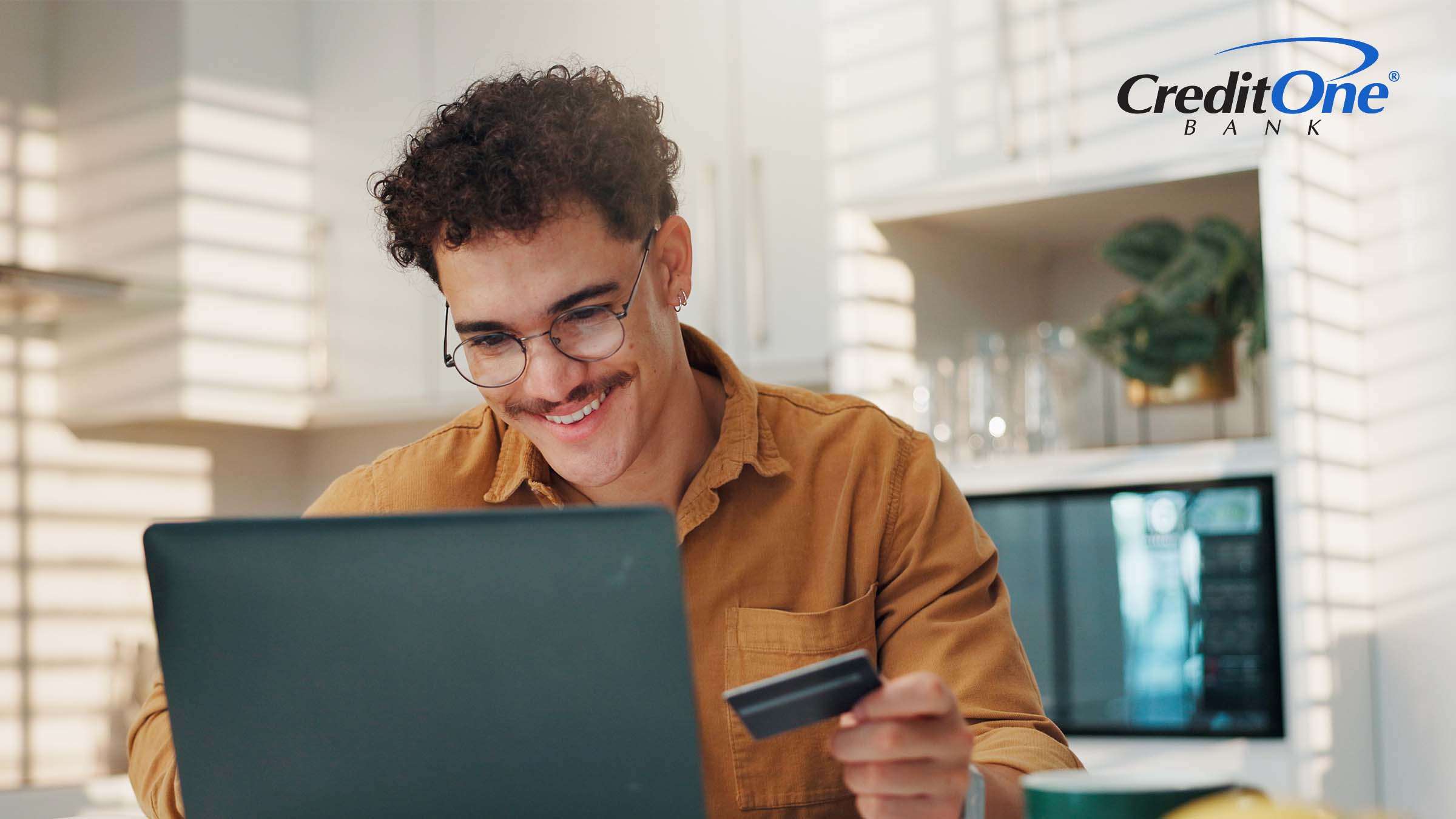 A man smiles as he looks at his laptop while holding a credit card. He may be making a credit card payment in his online bank or credit card account.